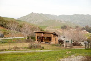A rustic wooden cabin with a porch, surrounded by a grassy landscape and hills in the background.