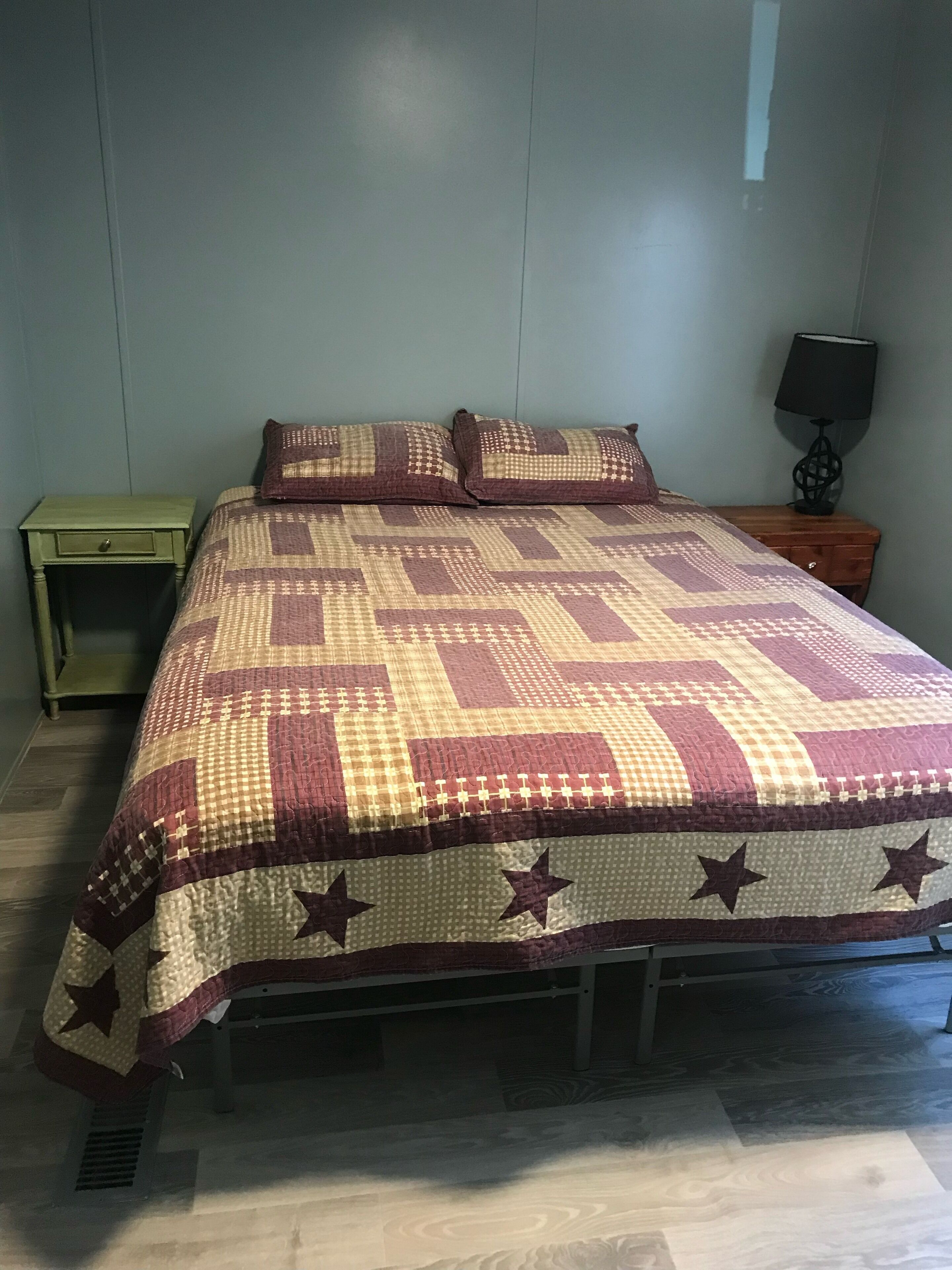 Un lit avec une courtepointe à motifs, deux oreillers, une table de chevet avec une lampe et un petit tiroir.