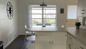 Una cocina con isla central, armarios blancos, una lámpara en el techo y una ventana con persianas.