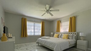 Un dormitorio con cama, mesitas de noche, ventilador de techo y ventana con cortinas.