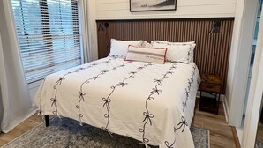 Un dormitorio con cama, ventana con persianas, unidad de aire acondicionado, un cuadro enmarcado en la pared y una mesita con una silla.