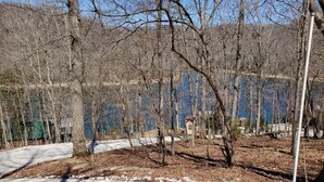 A snow-covered parking area with trees and a clear view of a lake and buildings in the background. - Hoot Owl Cabins -Incredible Lakefront Cabin PLUS a 2nd Tiny Home (Garfield)