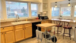 A kitchen with wooden cabinets, a granite countertop, a coffee maker, a microwave, a sink, a wooden table with stools, and a window with blinds.