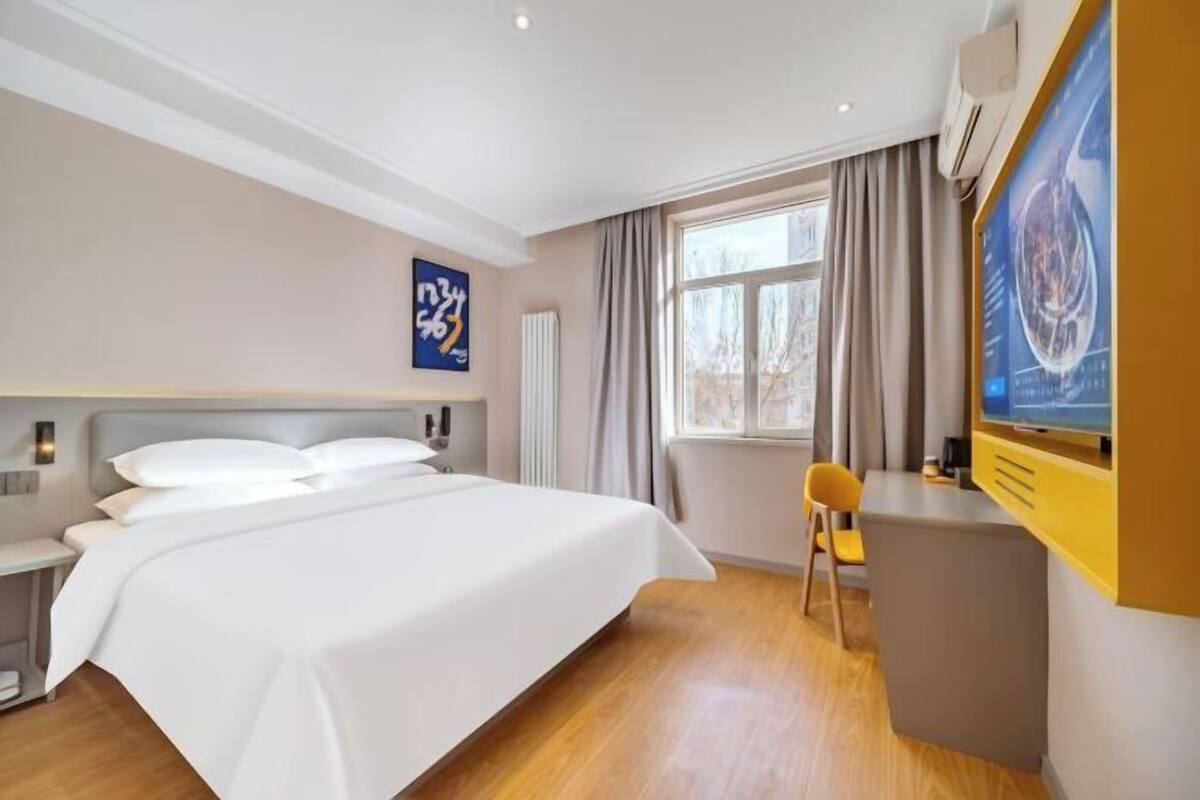 A hotel room with a bed, a desk, a chair, a window with curtains, and artwork on the wall.