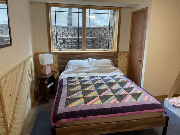 Quarto com cama de madeira, janela com vista para degraus de pedra, mesa de cabeceira com luminária e porta de madeira.
