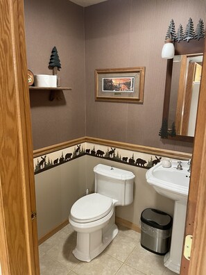Une salle de bain avec des toilettes, un lavabo et une poubelle. Un tableau encadré est accroché au mur, et une étagère contient un petit arbre et une bouteille.