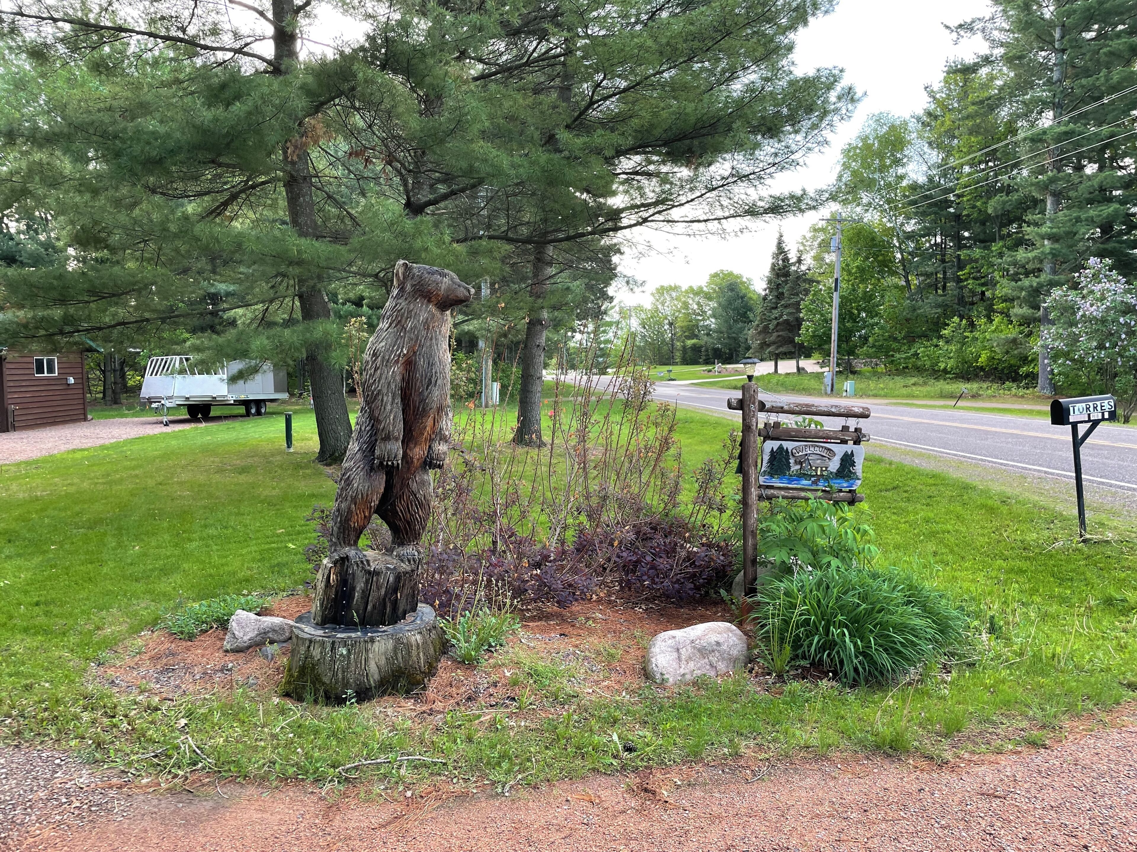 A rustic wooden bear sculpture in a grassy area with a welcome sign and mailbox.