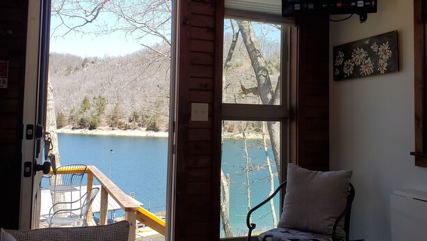 Interior acogedor de una cabaña con un gran ventanal que da a un lago, una zona de estar confortable y un televisor montado en la pared.