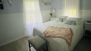 Un dormitorio con cama, ventilador de techo, banca y dos ventanas con cortinas de encaje.