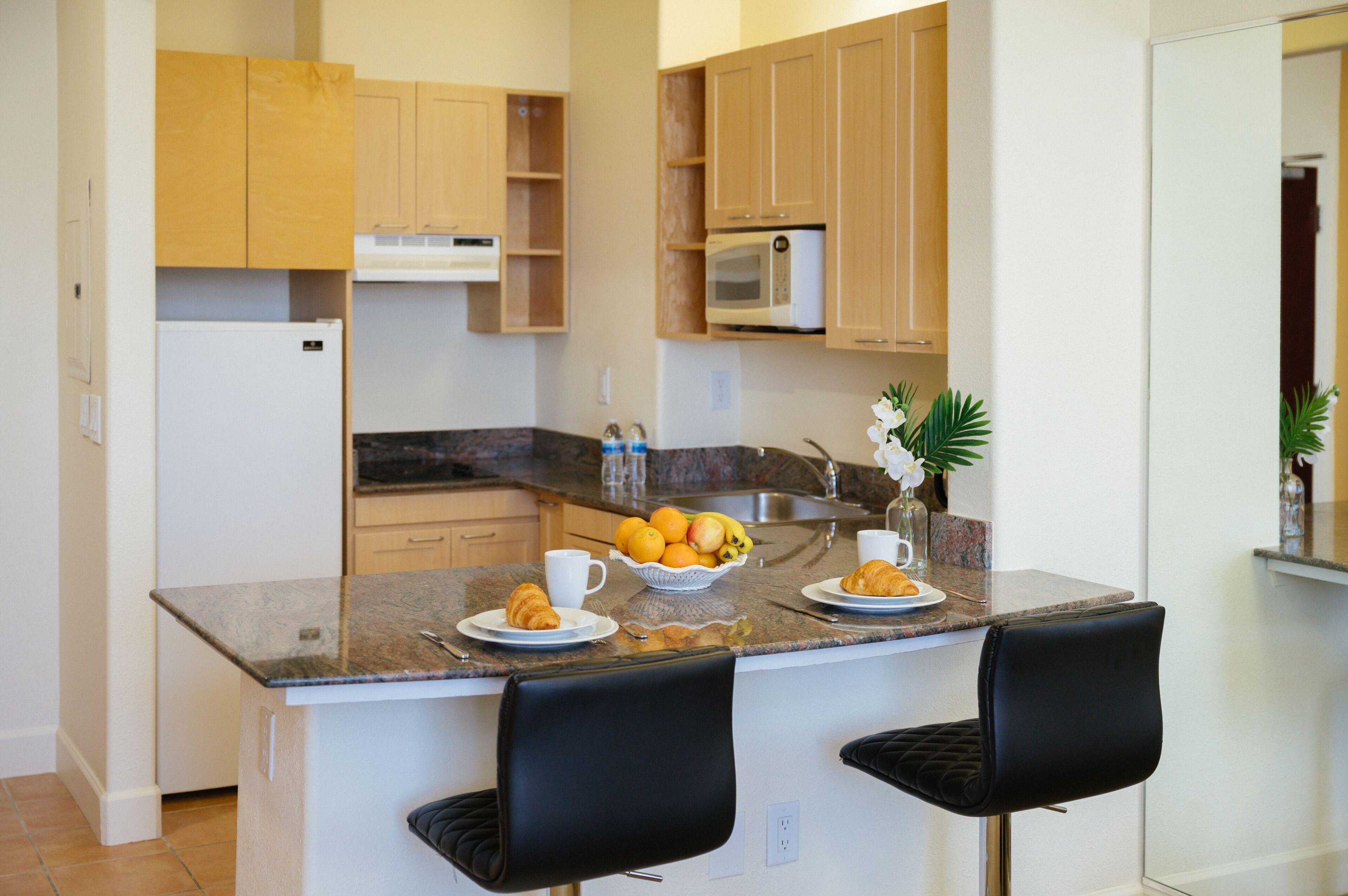 Een keuken met houten kastjes, een magnetron en een granieten aanrecht. Er staan twee zwarte stoelen en een schaal met fruit op het aanrecht.