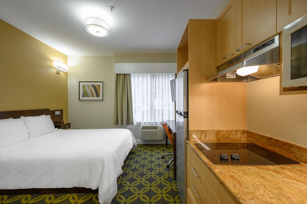 A hotel room with a bed, a desk, a kitchenette, and a window with curtains.
