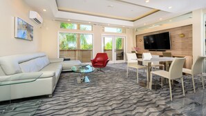 A modern living room with a glass coffee table, a red chair, a white sofa, and a dining area with a TV and wooden panel wall. - Studios Inn (San Jose)