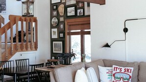 A cozy living room with a sofa, dining table, and chairs. A staircase with wooden railings leads to an upper level. - Cute kid-friendly condo next to Dollar Mountain (Sun Valley)