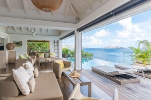 Una sala de estar moderna con un gran ventanal que da a una piscina y al mar. Se observan sofás, una mesa de centro y un comedor.