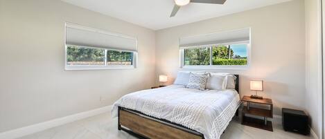 A bedroom with a bed, two bedside tables, a ceiling fan, and two windows with blinds.