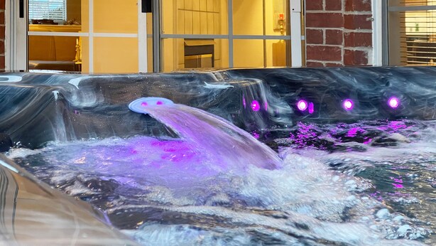 Una vasca idromassaggio con getti e illuminazione viola, sullo sfondo di un muro in mattoni e una finestra con persiane.