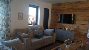 Una sala de estar con sofá, una mesa de centro, un televisor montado en una pared de madera y una ventana con vista a edificios.