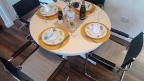 Una mesa redonda de comedor con cuatro sillas, cada una con un posavasos dorado y un plato blanco con diseño de hojas. Hay copas de vino, una botella de CAVA y un salero y pimentero sobre la mesa.