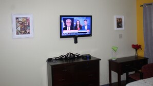 A hotel room with a television mounted on the wall, a wooden dresser, a desk with a lamp, and two framed artworks on the walls.