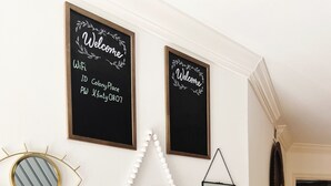 A wall with various decorative items including framed welcome signs, a mirror, a thermostat, and a decorative wall hanging.
