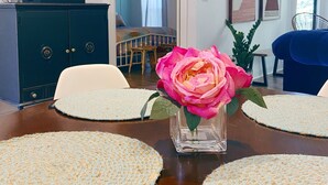 A dining table with placemats, a vase with a pink flower, and a view into a bedroom with a bed and curtains.