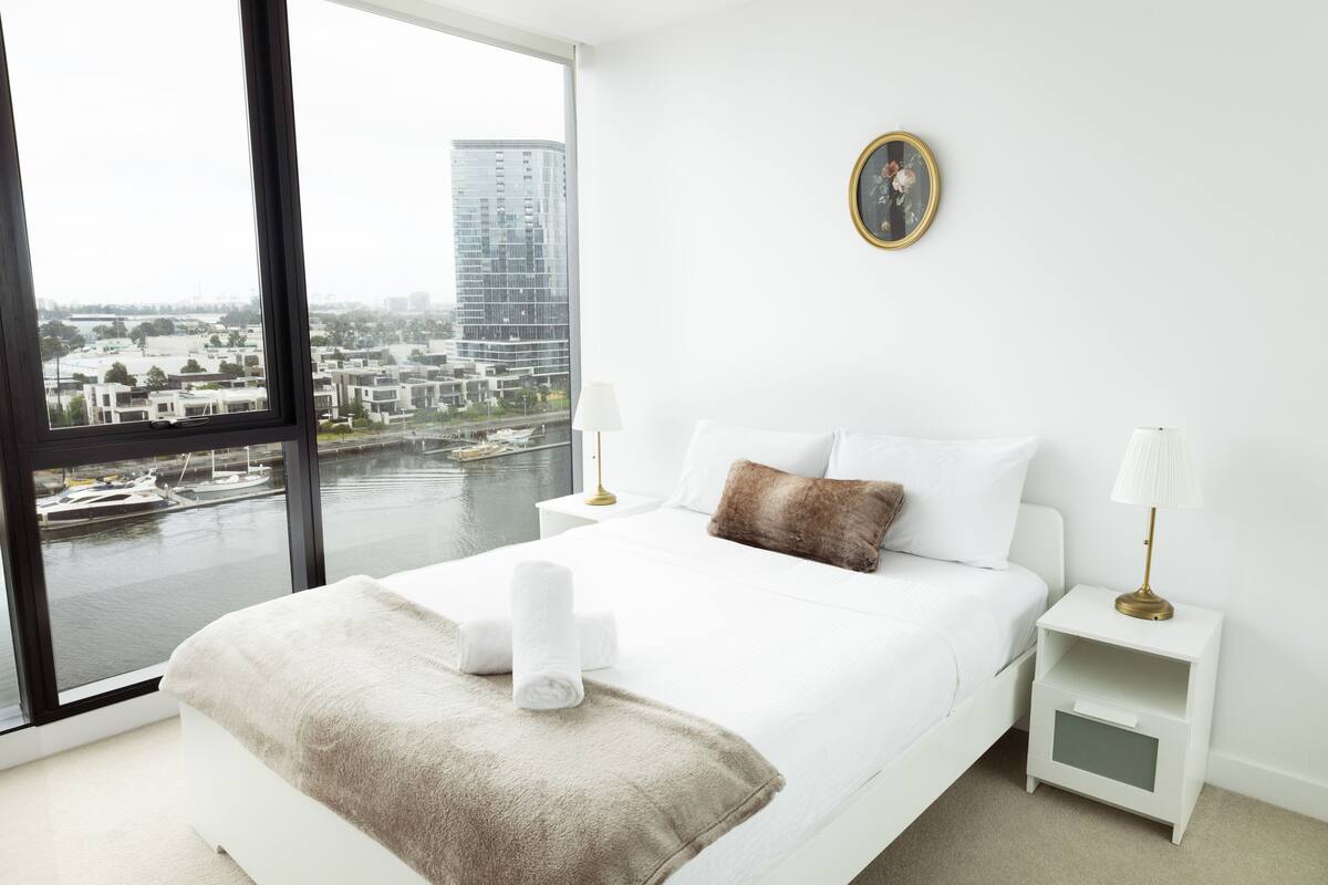 Una habitación de hotel con un ventanal grande, una cama con ropa blanca, un plaid marrón y una mesita de noche con lámpara.