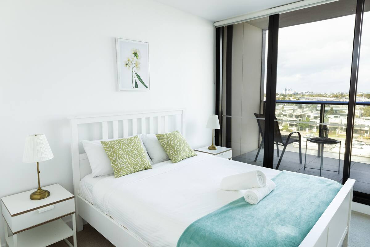 Habitación de hotel con una cama grande, ropa de cama blanca, una manta verde y vistas de la ciudad a través de amplias ventanas.