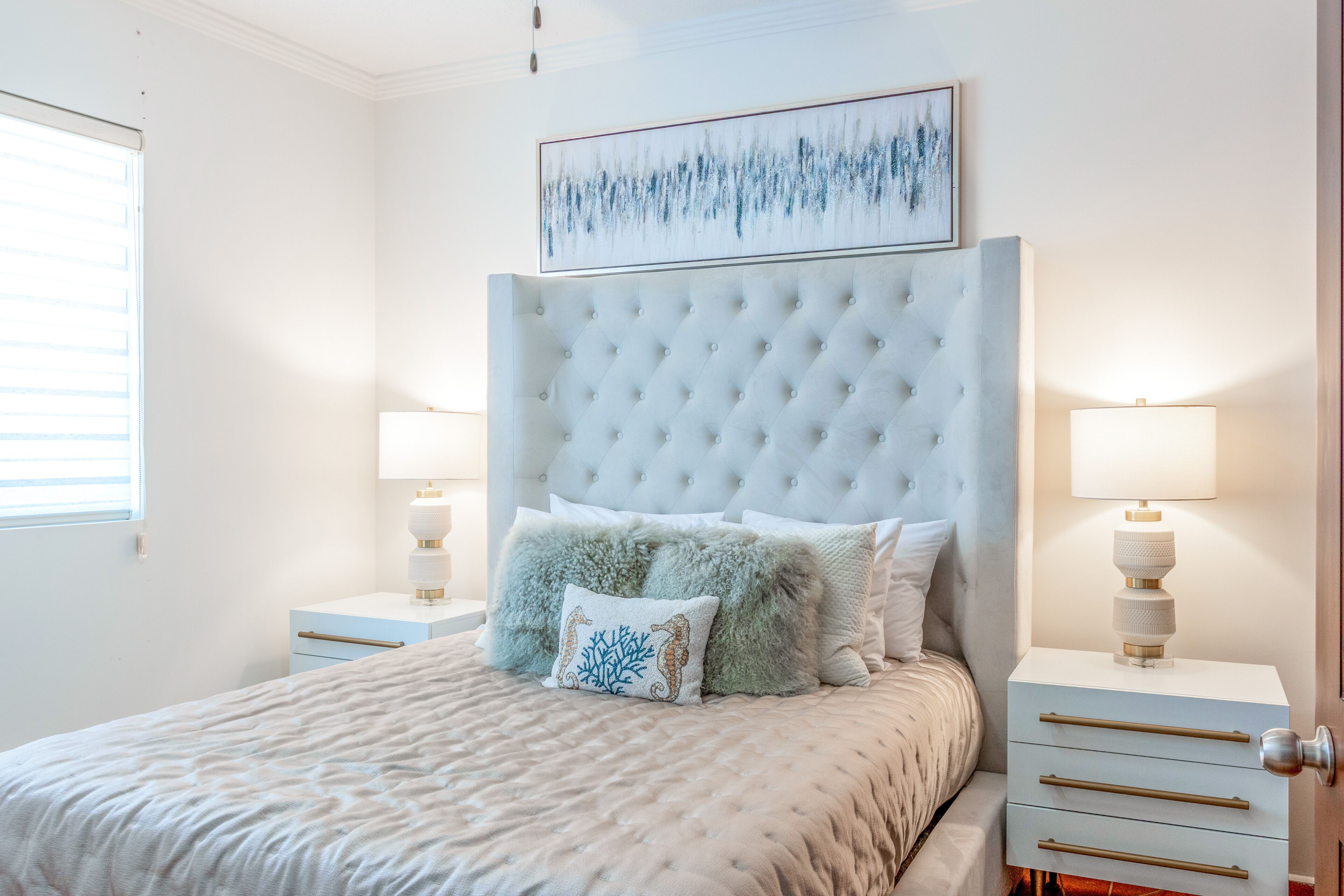 A bedroom with a tufted headboard, a bed with a textured bedspread, two bedside tables with lamps, and a framed artwork above the headboard.