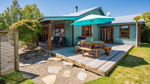 A wooden deck with a picnic table and a blue umbrella, a barbecue grill, and a cozy seating area with a sofa and cushions. - The Cottage on the Hill - Wānaka Holiday Home (Wanaka)