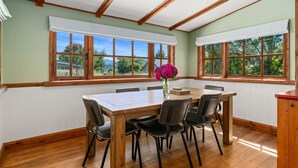 A dining area with a wooden table and chairs, large windows with white shutters, and a view of greenery outside. - The Cottage on the Hill - Wānaka Holiday Home (Wanaka)
