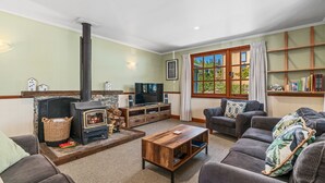 A cozy living room with a fireplace, comfortable seating, a TV, and a bookshelf. - The Cottage on the Hill - Wānaka Holiday Home (Wanaka)
