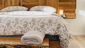 A bed with a wooden headboard, floral bedding, a bedside table with a lamp, and a small shelf with a plant.