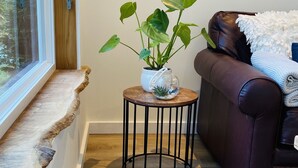 A cozy living room with a wooden floor, a brown leather sofa, a small round table with a plant, and a window with a wooden ledge.