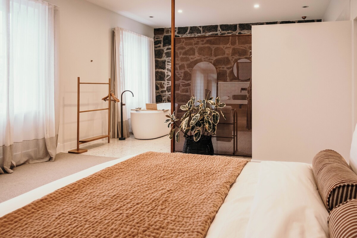 A bedroom with a large bed, a wooden ladder, a white bathtub, and a potted plant.
