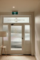 Entrada de hotel con puerta de vidrio esmerilado que muestra un logotipo y nombre, una mesita con lámpara, y un letrero de salida de emergencia verde.