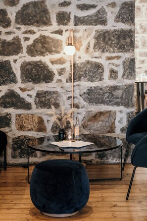 A room with a stone wall, a round glass table with a black metal base, a dark blue upholstered ottoman, and a tall floor lamp with a white shade.