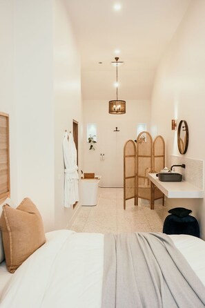 Quarto de hotel com cama, escrivaninha com pia, espelho e chuveiro.