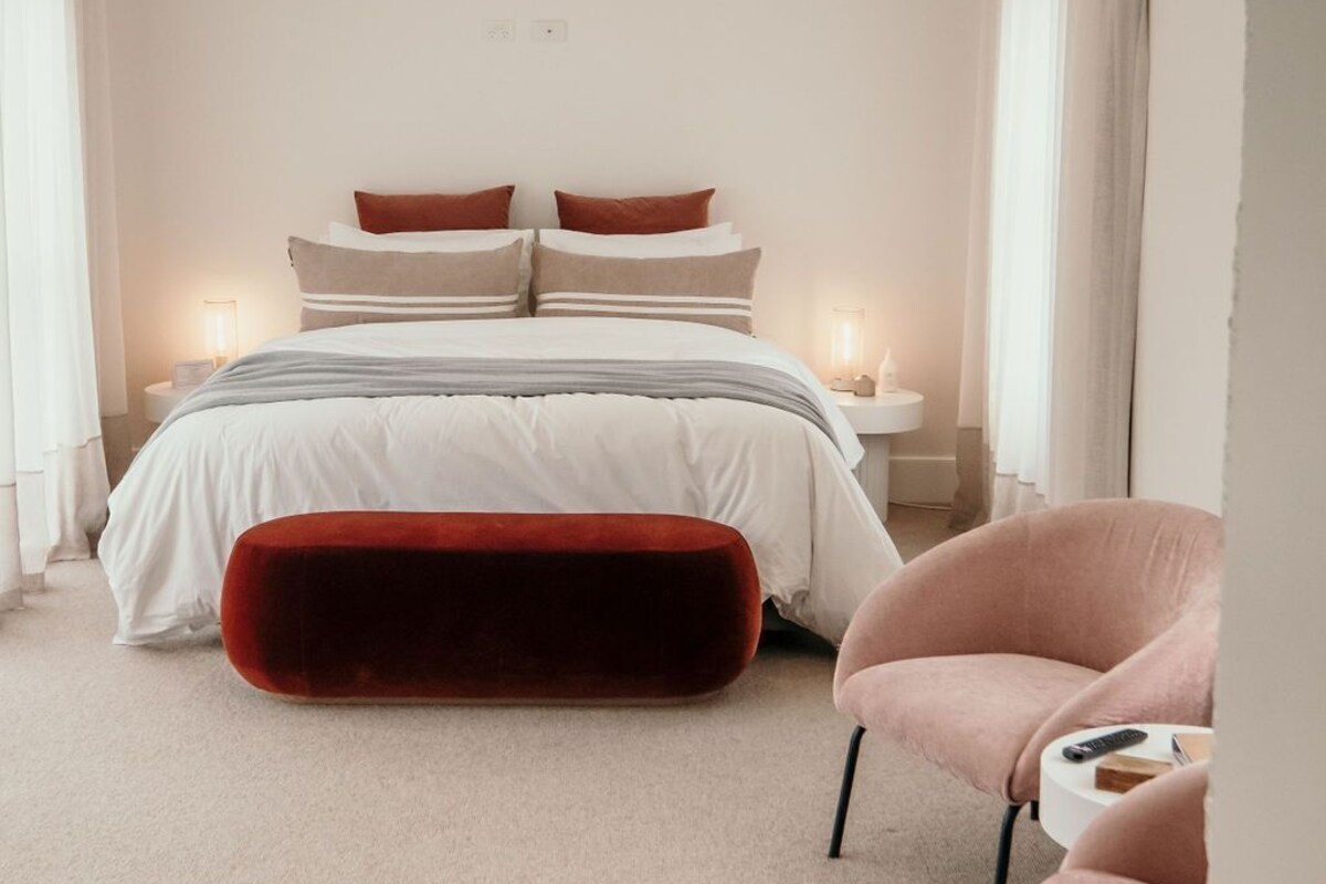 A bedroom with a bed, bedside tables, a red ottoman, and a pink chair.