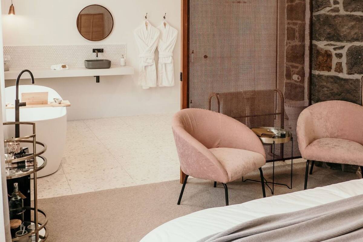 A modern bedroom with a bed, two pink chairs, a white vanity with a mirror, and a stone accent wall.