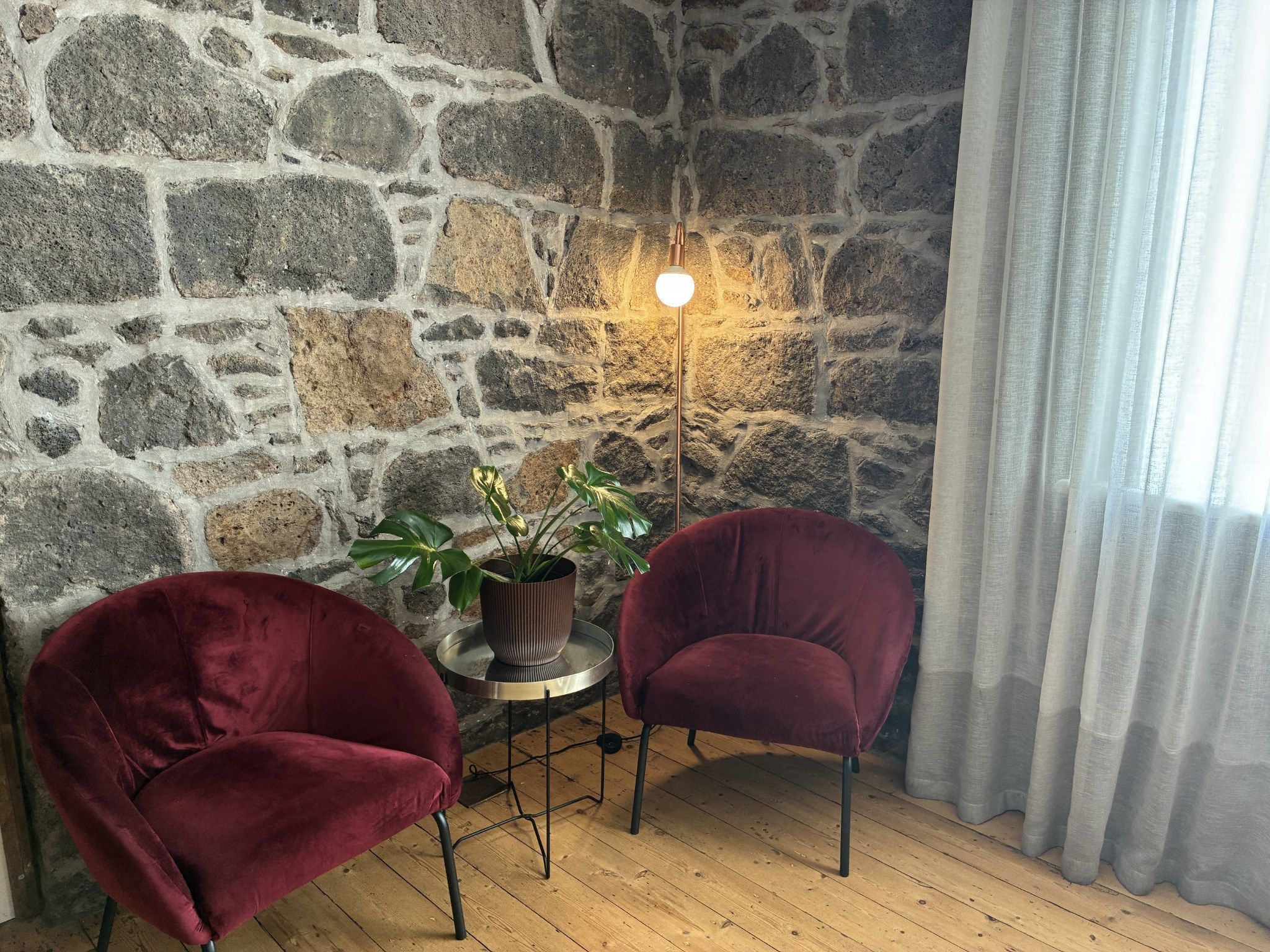 Una habitación con pared de piedra, dos sillas rojas, una mesita con planta en maceta y una lámpara de pie.