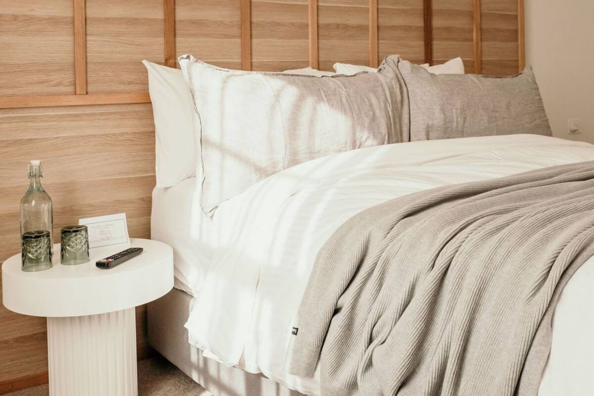 A bedroom with a wooden headboard, a round bedside table with a remote control, and a bed with a beige blanket.