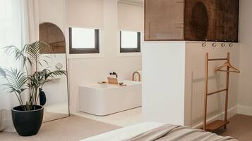 A modern bedroom with a large bed, a potted plant, a white bathtub, and a wooden drying rack.
