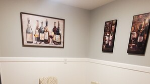 Un comedor con una mesa redonda y sillas estampadas, una lámpara de techo y tres cuadros con imágenes de botellas de vino en la pared.