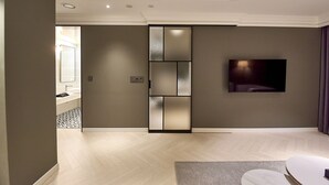Una sala moderna con un televisor de pantalla plana montado en la pared, un cerramiento de vidrio corredero que da al baño, y un suelo de madera con estampado tipo espiga.