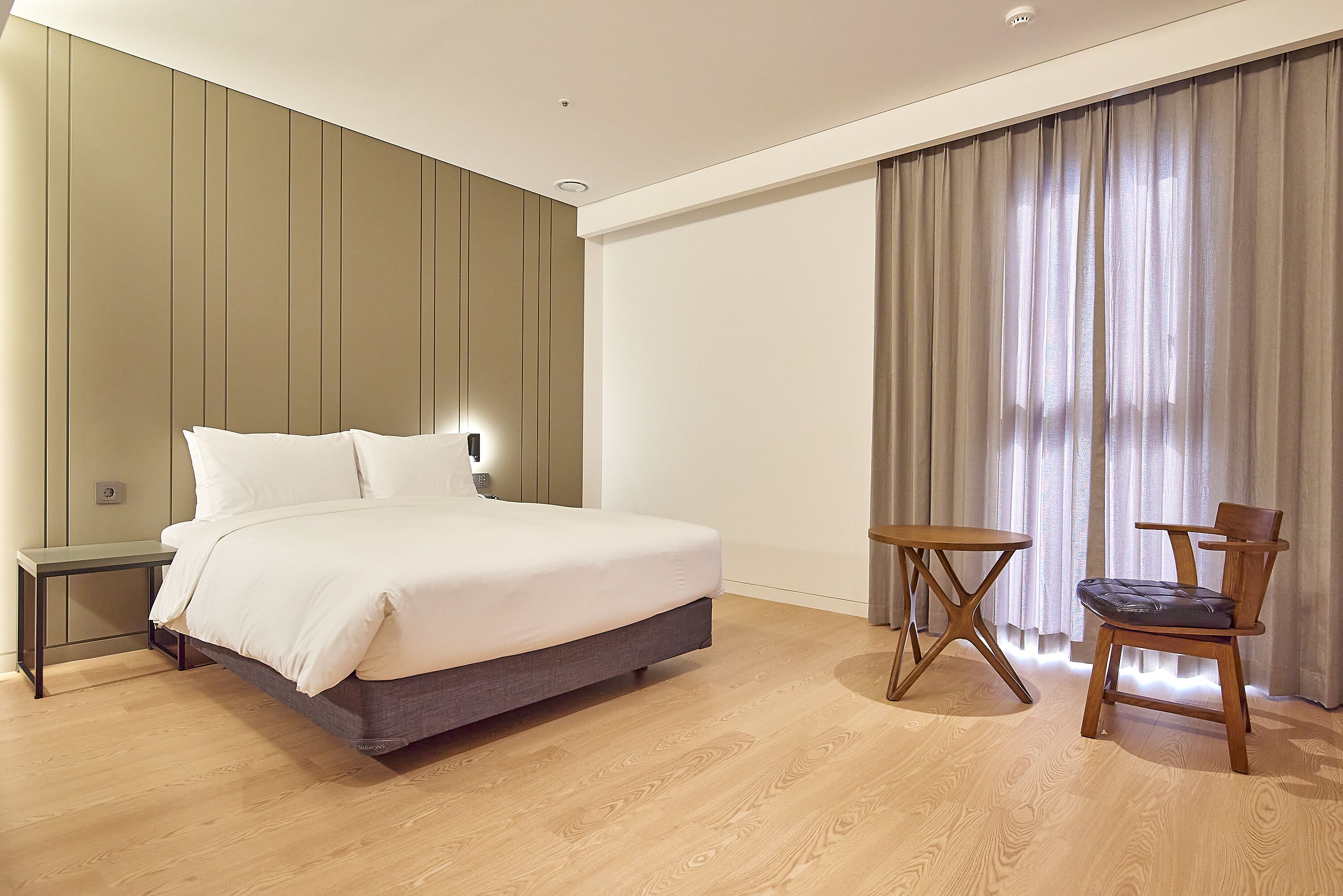 A modern hotel room with a large bed, a bedside table, a chair, and a small table.