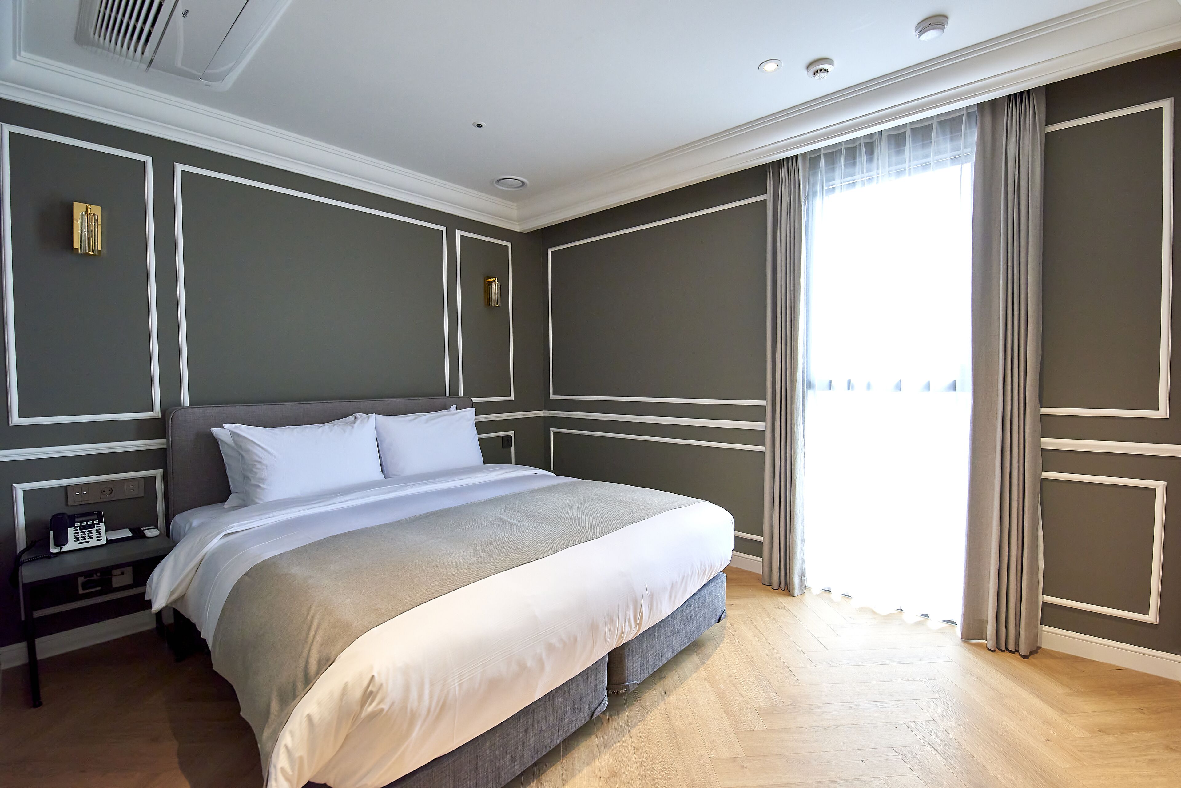A neatly made bed in a hotel room with a wooden floor, a nightstand with a telephone, and a window with curtains.