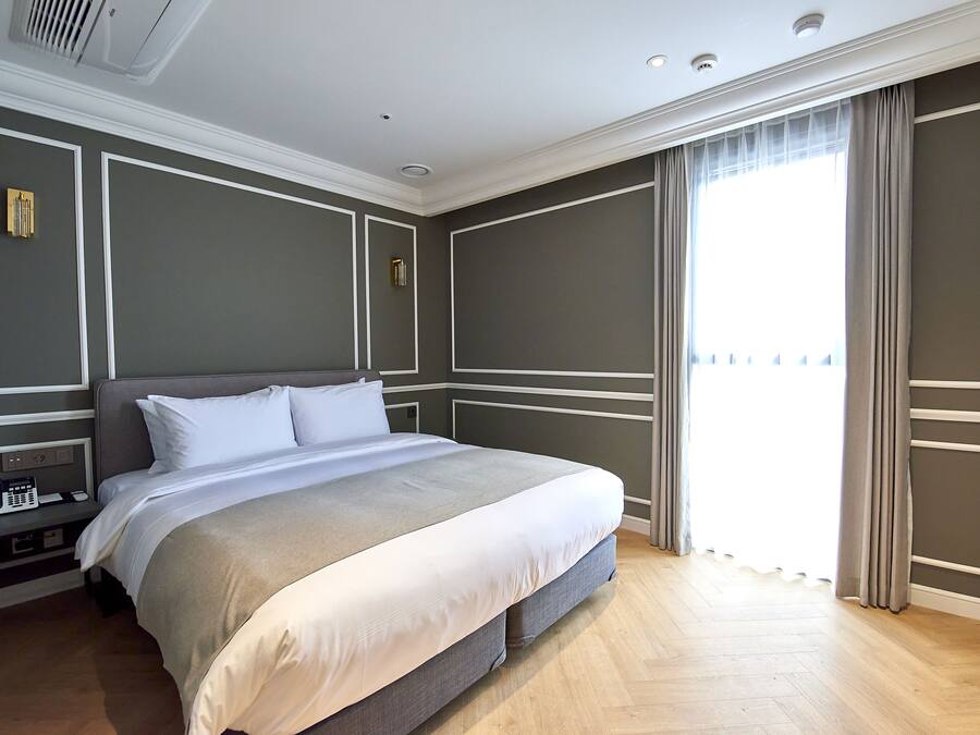 Una cama bien hecha en una habitación de hotel con suelo de madera, una mesita de noche con teléfono y una ventana con cortinas.