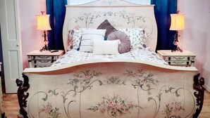 A bedroom with a floral-patterned bed, two bedside lamps, a chandelier, and blue curtains. - Historic Renovated Kemah Home just a short walk to the Kemah Boardwalk. (Kemah)