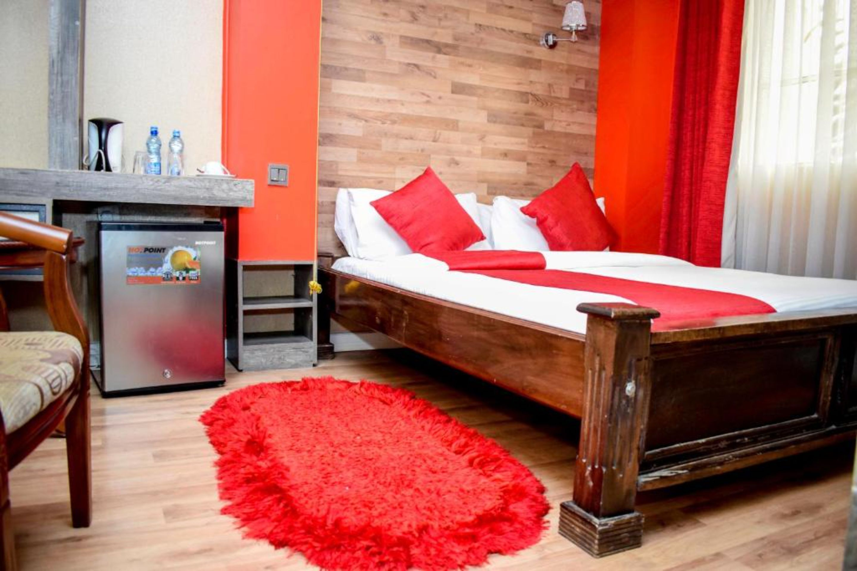 A hotel room with a wooden bed, red pillows, a red rug, a small refrigerator, and a wooden desk with a chair.