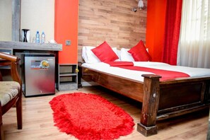 Habitación de hotel con una cama de madera, almohadas rojas, alfombra roja, un refrigerador pequeño y un escritorio de madera con una silla.